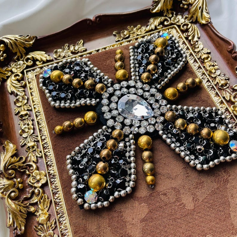 May include: A decorative butterfly design with black beads, gold accents, and a large clear gem. The butterfly is set on a brown fabric background within a decorative, ornate, brown and gold framed tray.