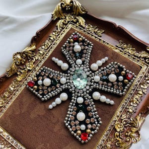 May include: A decorative cross with a central green gemstone, surrounded by pearls, red and clear crystals, and silver-colored beads. The cross is set on a brown velvet background within a decorative, ornate frame with gold accents.