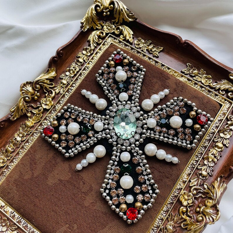 May include: A decorative cross with a central green gemstone, surrounded by pearls, red and clear crystals, and silver-colored beads. The cross is set on a brown velvet background within a decorative, ornate frame with gold accents.