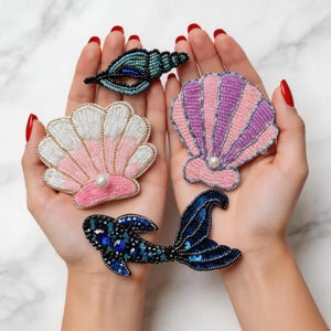Könnte beinhalten: Eine Sammlung von Perlenbroschen in Form von Muscheln und einer Meerjungfrauenflosse. Die Broschen zeigen rosa, weiße und lila Muscheln, eine blau-schwarze Meerjungfrauenflosse und eine türkisblaue Muschel, jeweils mit einer Perle.