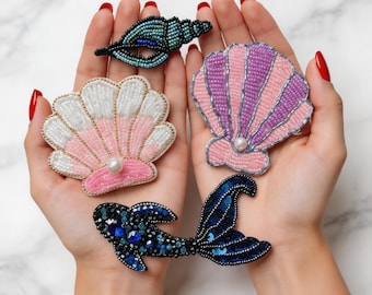 May include: A collection of beaded seashell and mermaid tail brooches. The brooches feature pink, white, and purple shells, a blue and black mermaid tail, and a teal seashell. Each brooch has a pearl accent.