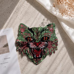 May include: A cat-shaped patch covered in green, pink, and black sequins. The design features a snarling feline face with red eyes, a red nose, and an open mouth showing teeth. The sequins create a textured, shimmering surface across the entire shape.