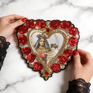 May include: A heart-shaped patch featuring a central image of a crowned figure holding a child, surrounded by a border of red and gold sequins. The patch has a black fabric backing and a scalloped edge, with intricate sequin detailing throughout.