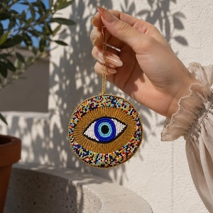 May include: A circular beaded ornament featuring a central eye motif with a black pupil, white iris, and blue outer ring. The background is gold, surrounded by a border of multicoloured beads. A thin gold chain is attached to the top for hanging.