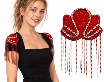 Puede incluir: Un par de hombreras rojas con diseño festoneado, adornadas con cristales transparentes y largos flecos de cuentas rojas colgantes. Las hombreras están sujetas a los hombros de un vestido negro con escote cuadrado.