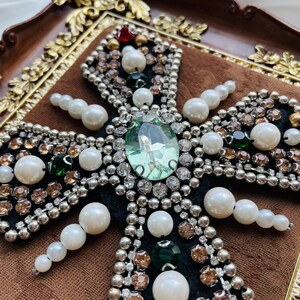 May include: Close-up of a decorative cross with a large, light green gemstone at the center. The cross is embellished with pearls, clear and amber-colored rhinestones, and smaller green and red gems. The cross is set against a dark background.