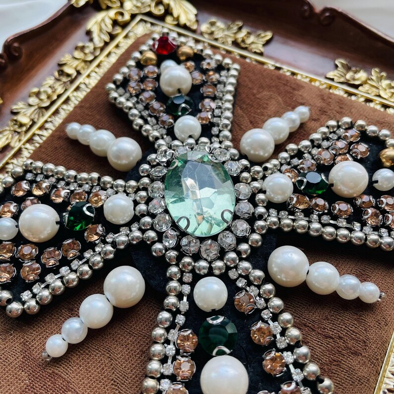 May include: Close-up of a decorative cross with a large, light green gemstone at the center. The cross is embellished with pearls, clear and amber-colored rhinestones, and smaller green and red gems. The cross is set against a dark background.