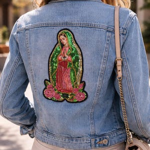 May include: Light blue denim jacket with a colorful embroidered patch of a religious figure on the back. The patch features a green and red design with pink roses at the bottom. The jacket has a classic design with button closures.
