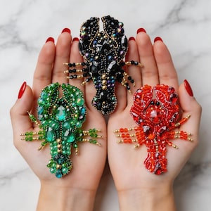 Puede incluir: Tres broches de langosta con cuentas en verde, negro y rojo. La langosta verde presenta gemas de color esmeralda, la negra tiene detalles iridiscentes y la roja está hecha de cuentas rojas. Cada broche tiene detalles dorados.