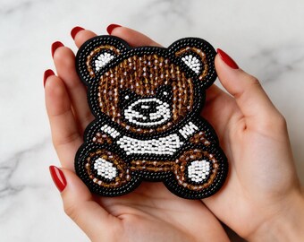 Puede incluir: Un parche de oso de peluche con cuentas marrones, blancas y negras. El oso tiene un contorno negro y se sostiene en manos abiertas. El cuerpo del oso es marrón, con detalles blancos en el pecho y la cara. El parche es un accesorio lindo y hecho a mano.