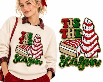 Puede incluir: Una sudadera color crema con un parche de chenilla texturizado con el texto TIS THE SEASON en rojo y verde. El diseño incluye un gráfico de una porción de pastel en capas y un árbol de Navidad con rayas rojas y blancas.