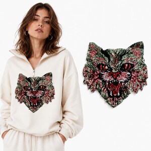 May include: A cream-coloured quarter-zip sweatshirt featuring a large, detailed cat face patch made of shimmering sequins in shades of olive green, pink, and black. The cat design displays an open mouth with visible teeth and sharp, focused eyes.