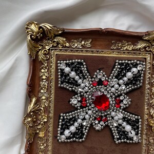 May include: A decorative framed artwork featuring a cross design. The cross is embellished with red, white, and clear gemstones, surrounded by black beads. The frame is brown with gold accents.