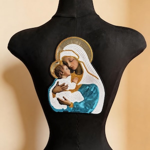 Può includere: Toppa religiosa raffigurante la Vergine Maria e Gesù bambino. L'immagine è incorniciata con paillettes dorate e bianche, con uno schema di colori blu e bianco. La toppa è applicata a un manichino nero.