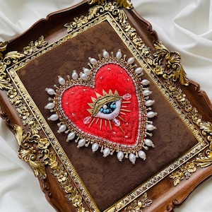 Könnte beinhalten: Gesticktes rotes Herz mit Augenmotiv, gerahmt in einem dekorativen Holzrahmen. Das Herz ist mit Perlen, Perlen und goldenen Akzenten verziert. Der Rahmen hat verzierte goldene Details.