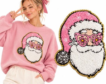 Puede incluir: Una sudadera rosa claro con un parche texturizado de Santa Claus con gafas rosas en forma de flor. El parche tiene un gorro rosa con borde blanco, una barba blanca y un pompón negro con lentejuelas plateadas, todo bordeado en oro.