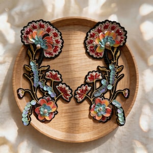 Può includere: Due toppe floreali ricamate con contorno nero, con paillettes e perline rosse, arancioni e blu. Le toppe sono su un piatto di legno. I fiori hanno una varietà di colori e trame.