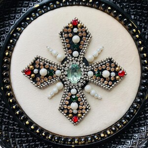May include: A decorative cross with a black frame. The cross is embellished with a variety of gemstones, including red, green, and clear crystals, and white pearls. The center features a large, oval-shaped, light green stone.