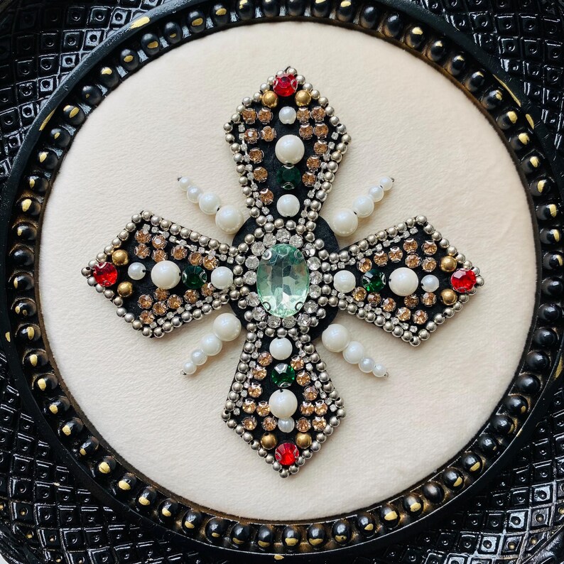 May include: A decorative cross with a black frame. The cross is embellished with a variety of gemstones, including red, green, and clear crystals, and white pearls. The center features a large, oval-shaped, light green stone.