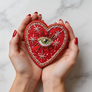 Puede incluir: Un parche bordado en forma de corazón rojo con un diseño de ojo. El parche está decorado con cuentas doradas, plateadas y blancas y patrones de estrellas. El parche se sostiene en manos abiertas.