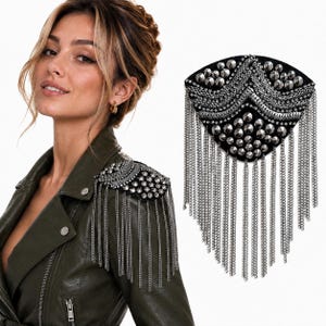 Könnte beinhalten: Eine schwarze Schulterepaulette mit silberfarbenen Metallnieten und mehreren Reihen herabhängender silberner Kettenfransen. Das dreieckige Accessoire ist für das Tragen auf der Schulter einer Jacke konzipiert und sorgt für Textur.