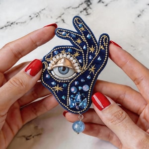 May include: A navy blue hand-shaped brooch with an eye design. The brooch features gold embroidery, blue crystals, and a dangling blue bead. The eye has a gold lid and a light blue iris. The brooch is held by hands with red nail polish.