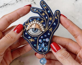 Puede incluir: Broche en forma de mano azul marino con diseño de ojo. El broche presenta bordados dorados, cristales azules y una cuenta azul colgante. El ojo tiene una tapa dorada y un iris azul claro.