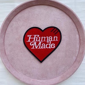 Pode incluir: Um patch vermelho em forma de coração com as palavras "Human Made" em escrita branca. O patch tem uma borda preta e está sobre um fundo circular rosa claro. O patch tem três linhas pretas no lado superior direito.