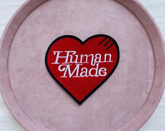 Puede incluir: Un parche rojo en forma de corazón con las palabras "Human Made" en escritura blanca. El parche tiene un borde negro y está sobre un fondo circular rosa claro. El parche tiene tres líneas negras en la parte superior derecha.
