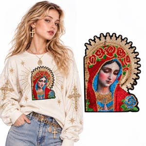 May include: A cream-coloured long-sleeve sweatshirt featuring a sequined patch of the Virgin Mary with a red head covering, blue garment, and gold halo. The sleeves and chest are decorated with gold embroidered cross and star motifs.