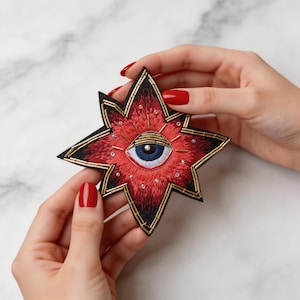 May include: Embroidered star-shaped patch with a red and black design. The patch features an eye in the center with a blue iris, surrounded by gold and white accents. The patch is held between two hands with red nail polish.