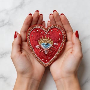 May include: A red heart-shaped embroidered patch with a gold crown and an eye design. The patch is embellished with gold beads and clear crystals. The background is a marble surface.