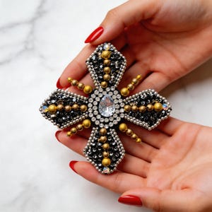 May include: A decorative cross-shaped brooch with a central teardrop-shaped clear crystal. The brooch features black, gold, and silver beads and crystals. The design is symmetrical and ornate, with a vintage or antique aesthetic.