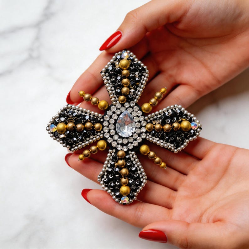 May include: A decorative cross-shaped brooch with a central teardrop-shaped clear crystal. The brooch features black, gold, and silver beads and crystals. The design is symmetrical and ornate, with a vintage or antique aesthetic.