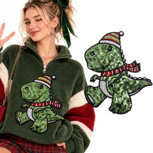 Può includere: Un maglione in pile verde scuro con mezza zip e una toppa a forma di dinosauro con paillettes. Il dinosauro è verde con la pancia bianca e indossa un berretto invernale e una sciarpa a righe rosse, bianche e verdi. La toppa è mostrata anche separatamente.