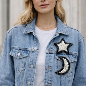 May include: A light blue denim jacket with a white t-shirt underneath. The jacket features two decorative patches: a black star with white beads and a black crescent moon with white beads. The jacket has silver buttons.