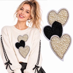 Puede incluir: Un suéter de manga larga color crema con tres aplicaciones en forma de corazón en el pecho, dos corazones cubiertos de perlas blancas y un corazón central de cuentas negras. Las mangas tienen detalles de cordones de cinta negra con lazos.