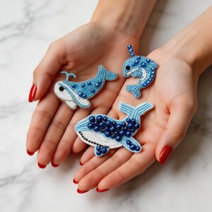 Peut inclure: Trois broches baleines perlées en nuances de bleu et de blanc. Les broches présentent des perles complexes, avec des perles bleu foncé accentuant les corps bleu clair. Une broche est un narval. Les broches sont tenues dans des mains ouvertes.