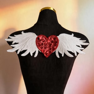 May include: A decorative applique featuring a red sequined heart with white embroidered wings. The heart is centered between the wings, creating a unique design. The applique is displayed on a black mannequin.