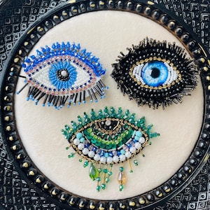 Puede incluir: Tres broches de ojo con cuentas en azul, negro y verde. El ojo azul tiene una base blanca con detalles azules y rosas. El ojo negro tiene un centro azul con cuentas doradas y negras. El ojo verde tiene cuentas verdes, azules y blancas con detalles colgantes.