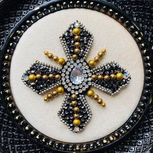 May include: A decorative cross with a central teardrop-shaped clear gem, surrounded by black, silver, and gold beads. The cross is set against a cream-colored background within a black, ornate frame.