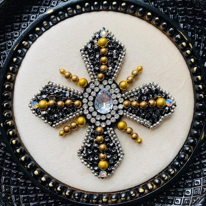 May include: A decorative cross with a central teardrop-shaped clear gem, surrounded by black, silver, and gold beads. The cross is set against a cream-colored background within a black, ornate frame.