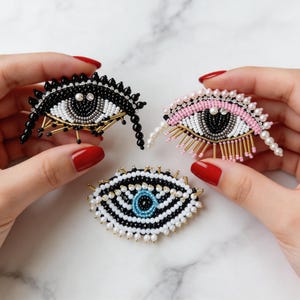 Op de afbeelding: Drie kralen oogbroches in zwart, roze en wit. Het zwarte oog heeft een zwarte kralenfranje en gouden accenten. Het roze oog heeft een roze bovenste wimperlijn en parelaccenten. Het witte oog heeft een blauw centrum.