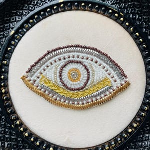 Op de afbeelding: Een gedetailleerd kralen oogontwerp in een zwarte lijst. Het oog is gemaakt met witte, zilveren, gouden en bruine kralen. De iris is een cirkel van bruine kralen met een gouden centrum. De lijst heeft een decoratief patroon.