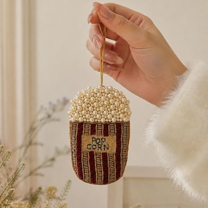 May include: A beaded popcorn ornament featuring a top section of white pearls and a bottom section with vertical stripes of dark red and gold beads. A rectangular patch in the center displays the embroidered black text POP CORN.