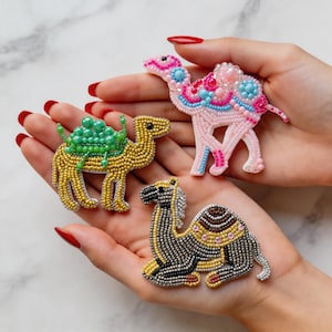 Puede incluir: Tres figuras de camellos con cuentas en amarillo, rosa y gris. El camello amarillo tiene una silla de montar verde, el camello rosa tiene detalles azules y rosas, y el camello gris está tumbado con detalles marrones y amarillos. Cada camello está intrincadamente adornado con cuentas.