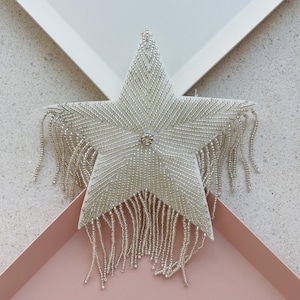 May include: A white star-shaped decoration with silver beadwork and fringe. The star has a central jewel and is set against a white and pink background. The star's design features a geometric pattern.