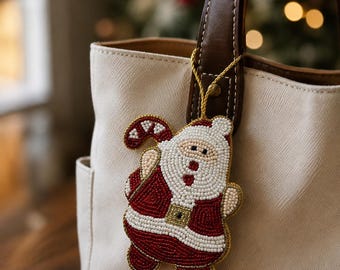 Puede incluir: Un adorno de cuentas con forma de Santa Claus y un bastón de caramelo, elaborado con cuentas de vidrio rojas, blancas y doradas. El adorno cuelga de un cordón dorado sujeto al asa de cuero marrón de un bolso de lona color crema.
