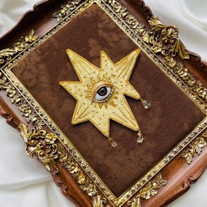 May include: A gold star-shaped embroidered patch with an eye in the centre, adorned with pearls and crystal teardrops. The patch is set on a brown velvet background within a decorative wooden frame with gold accents.