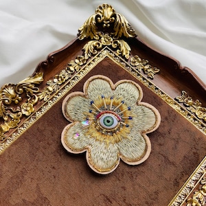 Puede incluir: Broche decorativo en forma de flor con un diseño de ojo en el centro. El broche presenta bordados dorados y beige, con un ojo verde, detalles rojos y adornos azules y dorados. Se encuentra sobre un fondo de terciopelo marrón dentro de un marco de madera decorativo.